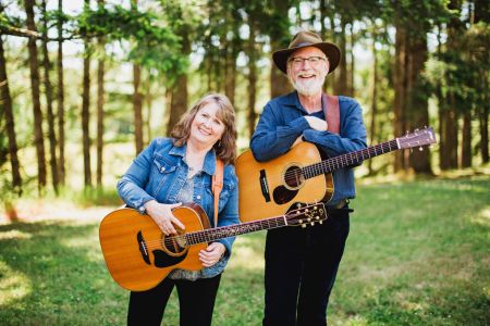 Mike and Carleen McCornack, both wearing guitars, standing in front of grass & trees.